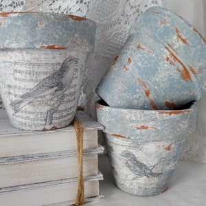 May include: Three blue and white terracotta flower pots with a distressed finish. The pots are decorated with a bird and musical notes design. The pots are stacked on top of each other.