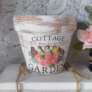 May include: White cottage garden planter with a distressed finish. The planter has a brown rim and features a floral design with birds and the text "Cottage Garden Bloom Where You Are Planted Est. 1901".