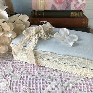 May include: A light blue box with a lace trim and a burlap ribbon tied in a bow. A single white flower is placed on top of the box. The box is sitting on a white lace tablecloth.
