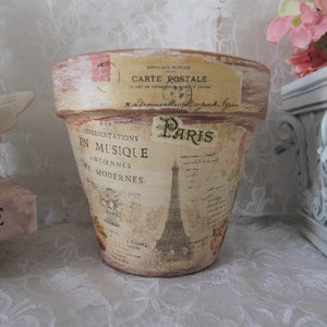 May include: A terracotta flower pot with a distressed white paint finish. The pot is decorated with a vintage Parisian theme, featuring an image of the Eiffel Tower, text in French, and a faded postcard. The text on the pot reads "Carte Postale" and "Des Representations en Musique Anciennes et Modernes."