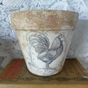 May include: A terracotta planter with a distressed white finish and a black and white rooster illustration. The planter has the text "Le Printemps" printed on the front.
