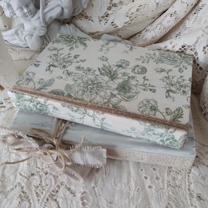 May include: A stack of decorative books with floral print covers in shades of green and white. The books are bound with a natural twine and ribbon. The background features lace and a small statue.