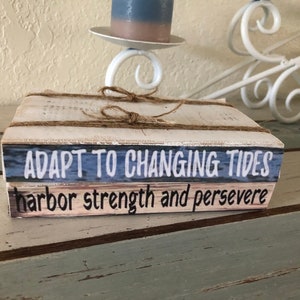 May include: Two stacked wooden blocks with a white background and blue and pink accents. The top block reads "Adapt to Changing Tides" and the bottom block reads "Harbor strength and persevere".