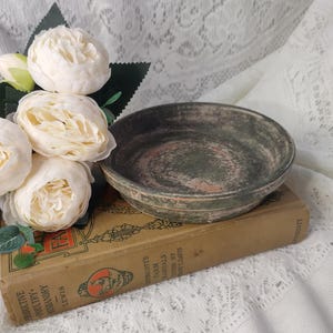 May include: A small, round, antique bowl with a distressed finish in shades of green, gray, and hints of red. The bowl rests on a vintage book with faded text and is accompanied by white peony flowers.