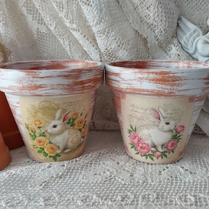 May include: Two terracotta flower pots with a distressed white paint finish. Each pot features a vintage-inspired design with a white rabbit and pink and yellow flowers.