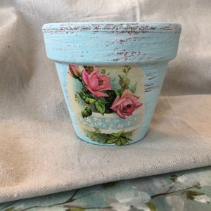 May include: A small, light blue terracotta flower pot with a distressed finish. The pot has a vintage floral design featuring pink roses and green leaves.