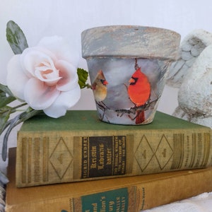May include: A terracotta flower pot with a distressed white finish. The pot is decorated with a winter scene featuring two red cardinals on a branch. The pot is sitting on top of two books.