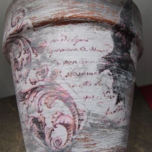 May include: A white ceramic planter with a distressed finish and a pink and black floral design. The planter has a wide mouth and a rounded bottom.