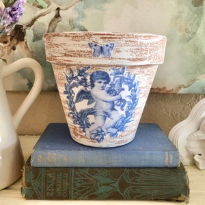 May include: White ceramic planter with a distressed finish, featuring a blue and white cherub design with a butterfly accent. The planter is sitting on top of a stack of books.