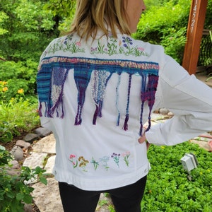 May include: White denim jacket with embroidered flowers and a colorful woven fringe trim. The fringe is made of blue, purple, and white yarn.