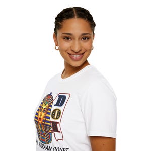 Peut inclure: T-shirt blanc avec un graphisme coloré représentant la tête d'un pharaon égyptien et les lettres "DOI" dans un design rectangulaire. Le texte "AL HARAAN COURT" est imprimé en dessous. Le t-shirt est en matière souple.