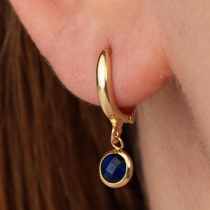 May include: A gold-coloured hoop earring with a dangling, round, deep blue gemstone. The earring is attached to an ear. The gemstone is faceted, catching the light. The earring's design is simple yet elegant.