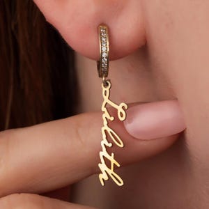 May include: Gold-coloured hoop earring with small clear stones set along the hoop. A gold pendant with the name "Lilith" in a cursive font hangs from the hoop. The earring is shown worn on an ear.