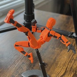 May include: An orange articulated action figure with black joint detailing, captured in a dynamic pose. The figure is mounted on a black stand, set against a blurred background with a tripod and wooden surface.