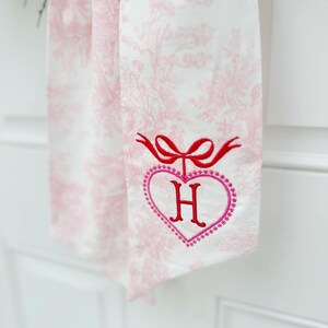 K&ouml;nnte beinhalten: Ein wei&szlig;es Stoffband mit einem rosafarbenen Blumenmuster. Das Band hat eine rote Schleife und ein rosafarbenes Herz mit dem Buchstaben "H" in der Mitte gestickt.