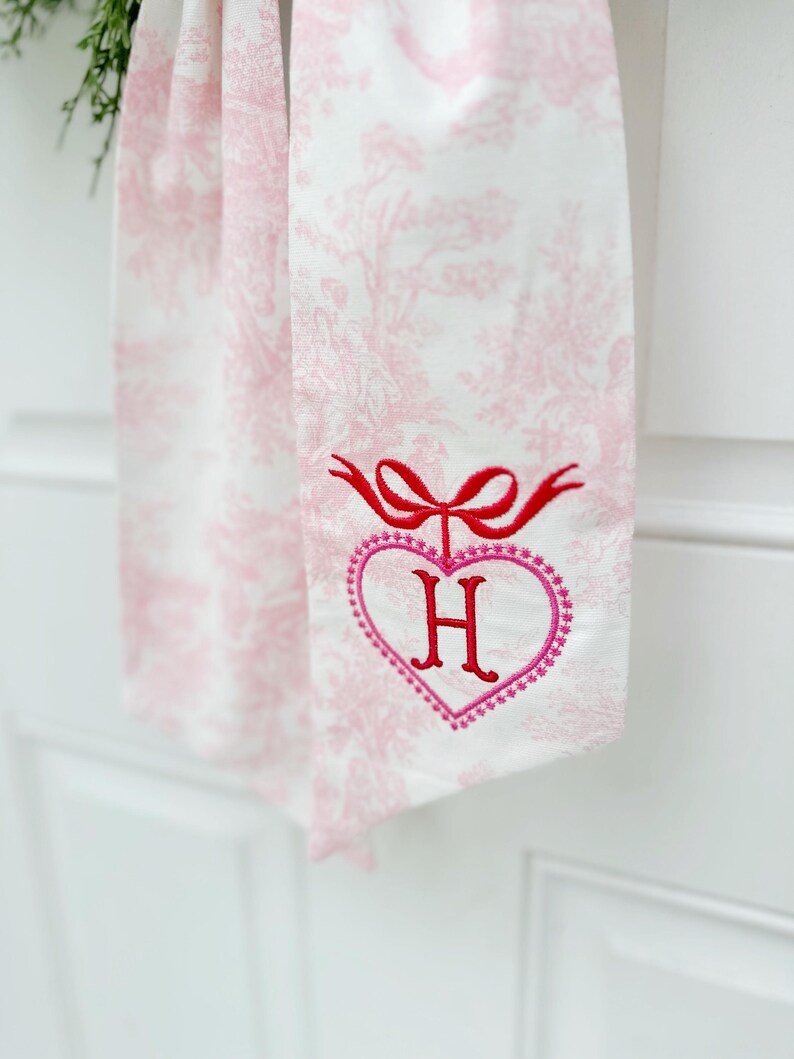 K&ouml;nnte beinhalten: Ein wei&szlig;es Stoffband mit einem rosafarbenen Blumenmuster. Das Band hat eine rote Schleife und ein rosafarbenes Herz mit dem Buchstaben "H" in der Mitte gestickt.