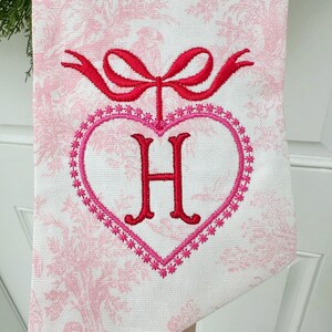 K&ouml;nnte beinhalten: Ein wei&szlig;es K&uuml;chentuch mit einem rosa Blumenmuster. Das Tuch hat eine rote gestickte Schleife und ein rosa Herz mit einem roten gestickten Buchstaben H in der Mitte.