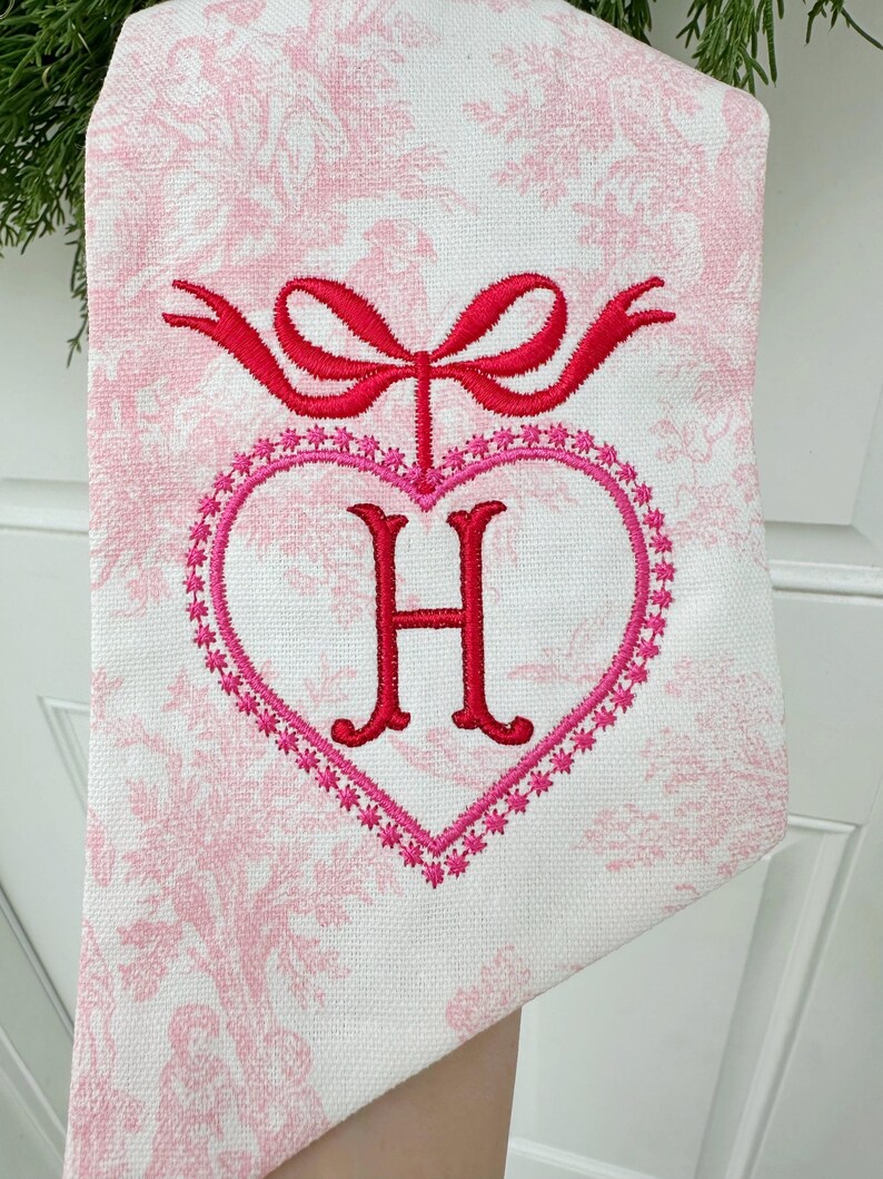 K&ouml;nnte beinhalten: Ein wei&szlig;es K&uuml;chentuch mit einem rosa Blumenmuster. Das Tuch hat eine rote gestickte Schleife und ein rosa Herz mit einem roten gestickten Buchstaben H in der Mitte.
