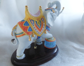 May include: A white porcelain elephant figurine with a colorful saddle and gold accents. The elephant is standing on a wooden base and is holding a small, blue and white patterned drum.