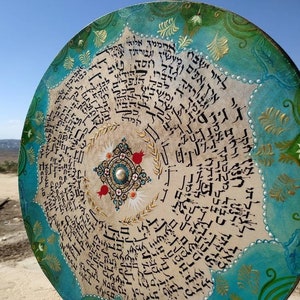 May include: A round, decorative plate with a turquoise and green border. The center of the plate is white with black Hebrew writing and a gold and white design. The plate is decorated with gold and white floral designs.