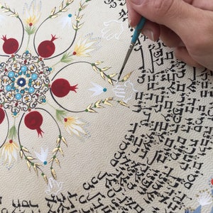 May include: A close-up of a hand painting a white design on a circular piece of parchment paper. The parchment is decorated with a central mandala design, red pomegranates, and gold leaf accents. The text surrounding the design is in Hebrew.