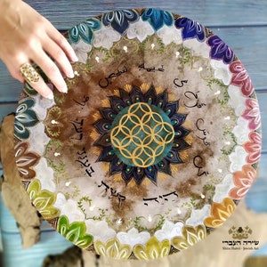 May include: A round wooden plate with a mandala design in the center. The mandala is painted in shades of blue, green, and gold. The plate is surrounded by a border of colorful flowers and leaves. The words "שירה העברי" are written in Hebrew at the bottom of the plate.