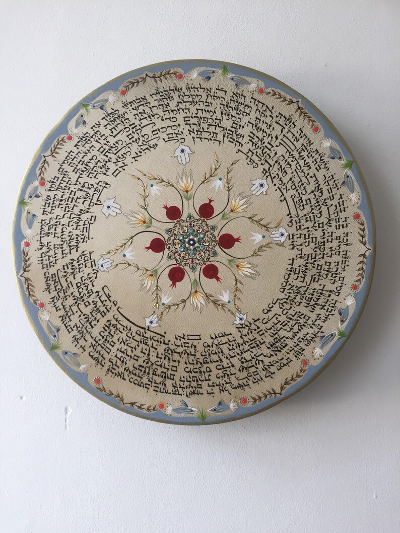 May include: A round, cream-colored plate with a blue border. The center of the plate features a floral design with pomegranates and a Hamsa hand. The plate is decorated with Hebrew text around the edge.