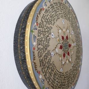 May include: A round, wooden drum with a beige surface and a blue border. The drum is decorated with a circular design of flowers, pomegranates, and Hebrew text. The drum is surrounded by a black and gold border.