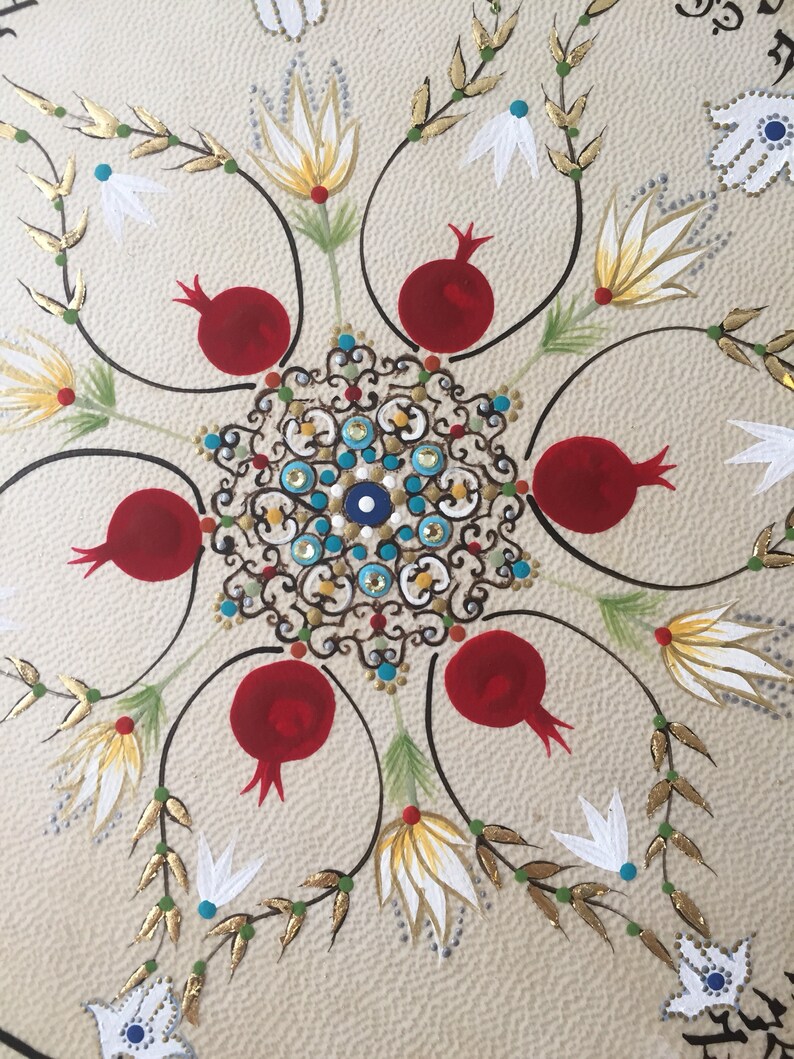 May include: A circular mandala design with a central eye motif surrounded by six red pomegranates. The pomegranates are surrounded by stylized white flowers with gold accents and green leaves.