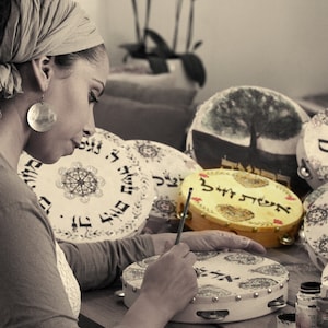 May include: A woman paints a round wooden tambourine with black and white designs. The tambourine is surrounded by other tambourines with different designs, including a tree, Hebrew text, and other patterns. The tambourines are on a table with paint bottles.
