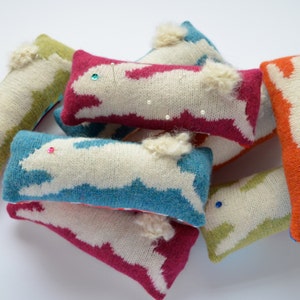 May include: A collection of colorful knitted bunny pincushions. The pincushions are made of soft, fluffy yarn and have a variety of colors, including pink, blue, green, and orange. Each pincushion has a white bunny design and a fluffy tail.
