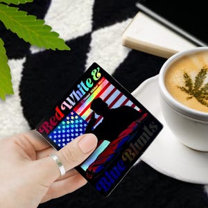 May include: A square sticker with the text "Red White & Blue Blunts" and an American flag design. The sticker features a silhouette of a person and a cannabis leaf design in the coffee.