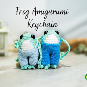 Puede incluir: Dos llaveros de rana de ganchillo. Las ranas son de color verde claro y blanco con petos azules. El texto "Frog Amigurumi Keychain" está en la parte superior de la imagen. El fondo está borroso con una linterna blanca y una cuerda.