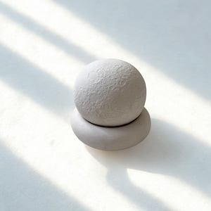 Może przedstawiać: Jasnoszary, kulisty przedmiot dekoracyjny o teksturowanej powierzchni przypominającej księżyc, spoczywający na pasującej, zaokrąglonej podstawie. Projekt jest minimalistyczny i nowoczesny, odpowiedni do wystroju wnętrz. Obraz jest oświetlony naturalnym światłem.