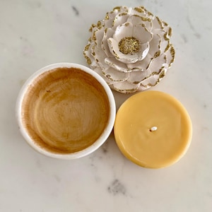 May include: A small, round, open bowl with a white exterior and a brown interior sits next to a yellow candle and a white flower with gold glitter accents. The candle has a white wick.
