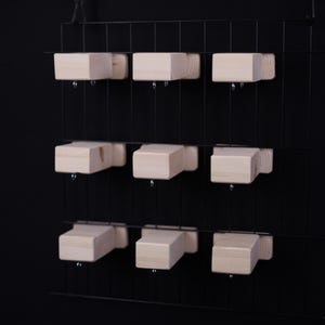 May include: A black metal grid with nine light-colored wooden blocks suspended from it. The blocks are evenly spaced and appear to be attached with small metal hooks. The background is a solid black.