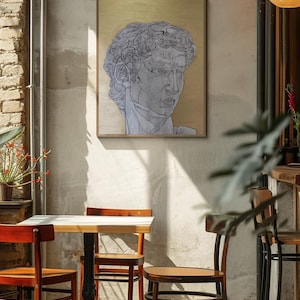 Puede incluir: Una obra de arte enmarcada que presenta un boceto a lápiz detallado de un busto clásico sobre un fondo dorado. La obra de arte se exhibe en un ambiente de cafetería, con una pequeña mesa y sillas en primer plano. La escena está iluminada por luz natural.