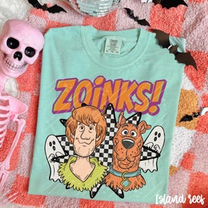May include: A light mint green t-shirt featuring a graphic of Shaggy and Scooby-Doo with the text ZOINKS! in orange and purple. The design includes a black and white checkered star shape and two white ghost illustrations on a crew neck shirt.