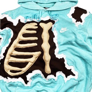 May include: A light blue Nike hoodie with a unique design. The hoodie features a brown patch with a bone-like design and white accents. The Nike logo is visible on the front. The hood and edges have a wavy, torn look.