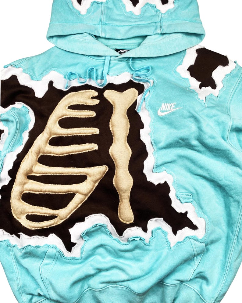 May include: A light blue Nike hoodie with a unique design. The hoodie features a brown patch with a bone-like design and white accents. The Nike logo is visible on the front. The hood and edges have a wavy, torn look.