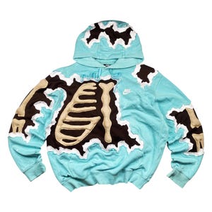 May include: A light blue hoodie with a unique skeleton design. The design features a brown and tan skeleton applique with white trim. The hoodie has a hood and a small Nike logo on the chest.