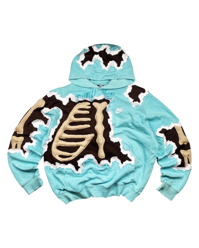 May include: A light blue hoodie with a unique skeleton design. The design features a brown and tan skeleton applique with white trim. The hoodie has a hood and a small Nike logo on the chest.