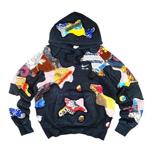 Puede incluir: Sudadera con capucha negra con diseños de patchwork coloridos. La sudadera con capucha tiene un bolsillo de canguro y una capucha. El patchwork incluye varios patrones y colores, con la palabra "AIR" impresa en el pecho.