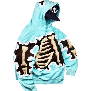 May include: A light blue hoodie with a brown and beige skeleton design. The hoodie has a torn edge design and a navy blue baseball cap with a white logo. The hoodie is made of a soft material.