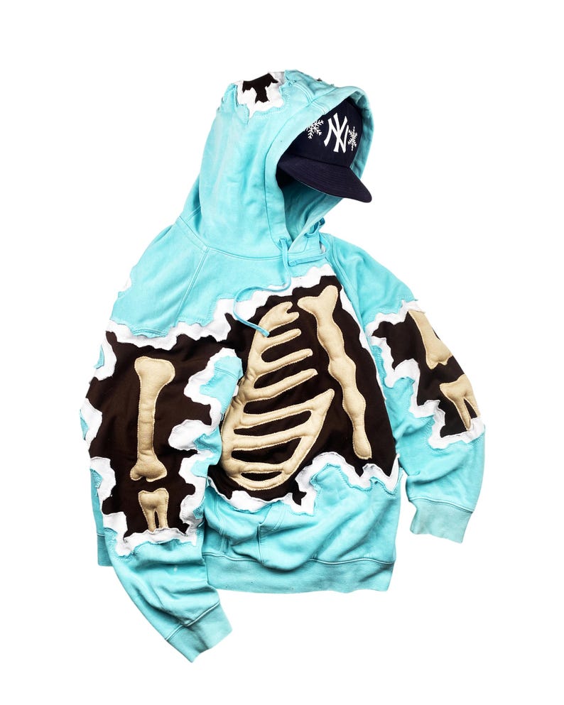 May include: A light blue hoodie with a brown and beige skeleton design. The hoodie has a torn edge design and a navy blue baseball cap with a white logo. The hoodie is made of a soft material.