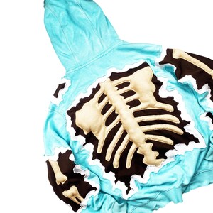 May include: A light blue hoodie with a skeleton design on the back. The skeleton is beige with a dark brown outline and white trim. The hoodie has a hood and long sleeves.