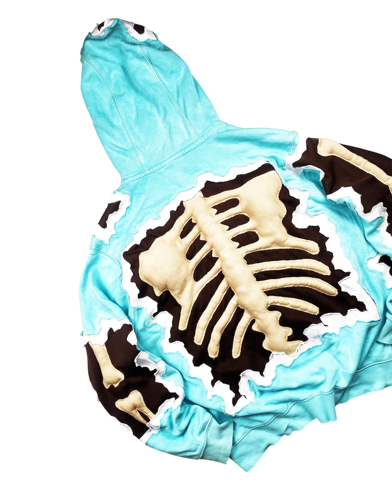 May include: A light blue hoodie with a skeleton design on the back. The skeleton is beige with a dark brown outline and white trim. The hoodie has a hood and long sleeves.