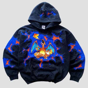 Op de afbeelding: Zwarte hoodie met een kleurrijk drakenontwerp, met oranje vlammen en blauwe accenten. Het ontwerp is geplaatst tegen een donkere achtergrond. De hoodie heeft een voorzak en een capuchon met trekkoord.