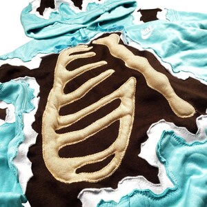 May include: A turquoise hoodie with a brown rib cage design. The rib cage is made of a tan material and is sewn onto the brown fabric. The hoodie has a white, wavy trim and a matching hood.