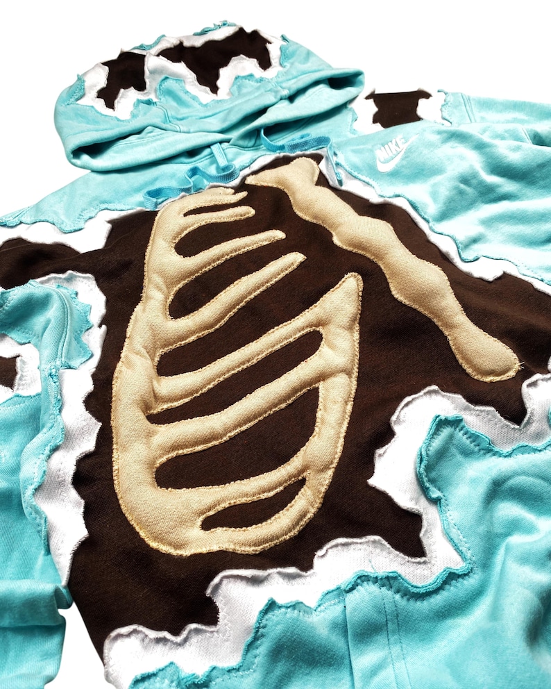 May include: A turquoise hoodie with a brown rib cage design. The rib cage is made of a tan material and is sewn onto the brown fabric. The hoodie has a white, wavy trim and a matching hood.